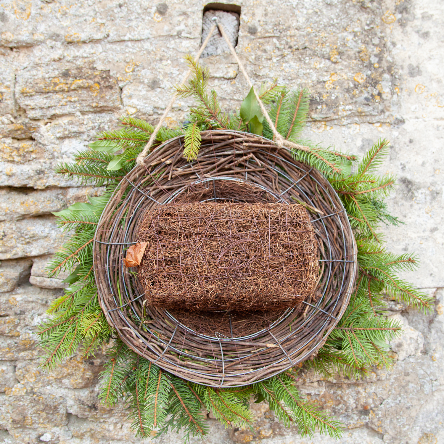 Back of wreath shown hung on a stone wall. Wreath is made of brushwood and foliage. Back of wreath shows rectangular nest box.
