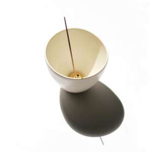 White porcelain curved pot shown with brass incense holder inside with incense stick inserted.