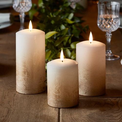 A trio of bronxe ombre led pillar candles positioned on a dark wood table with two wine glasses and greenery in the backround.