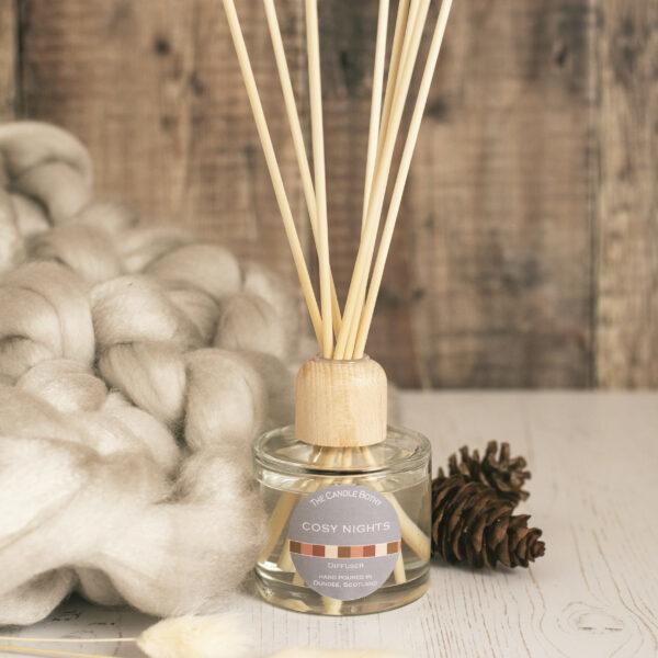 Cosy nights reed diffuser in between a grey merino wool chunky knit blanket and two pine cones.
