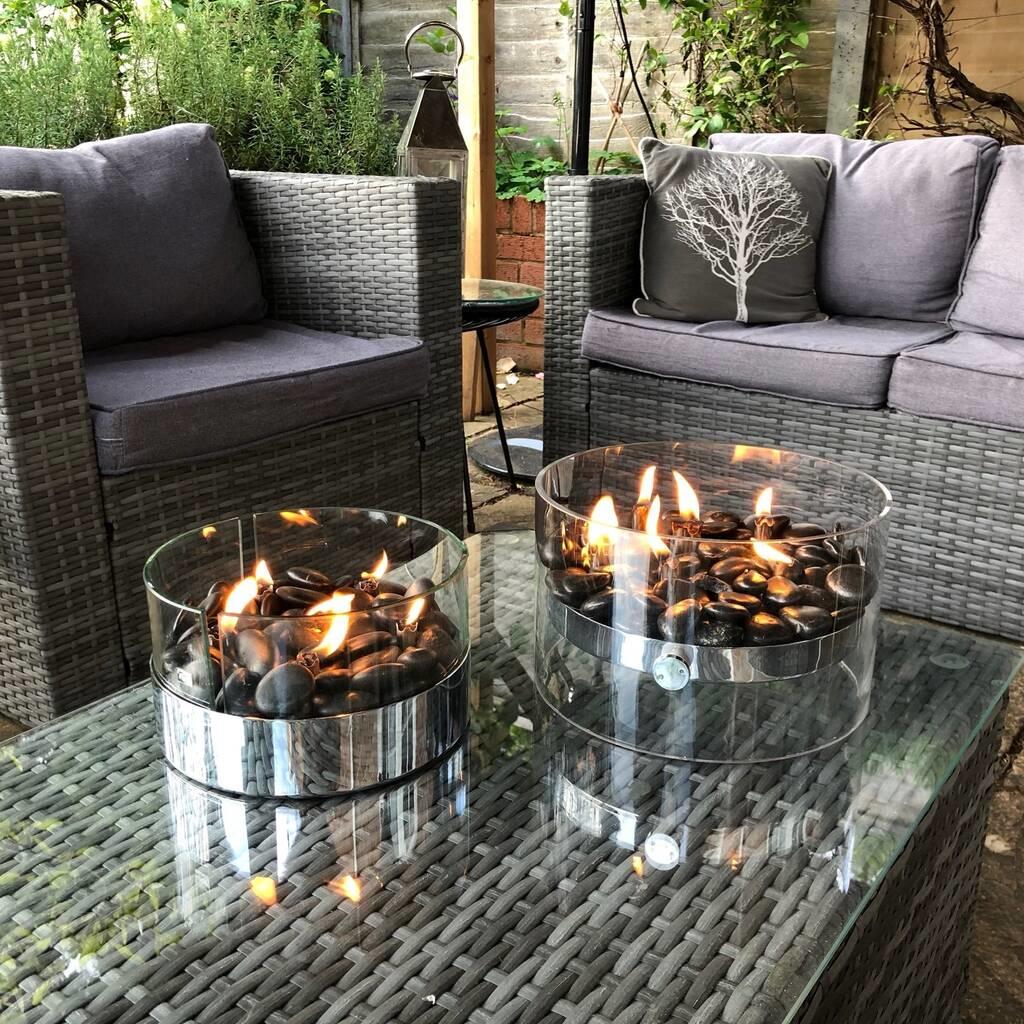 Two lit cylindrical glass tabletop fire pits on a grey outdoor wicker table, with wicker sofas and a tree design pillow.