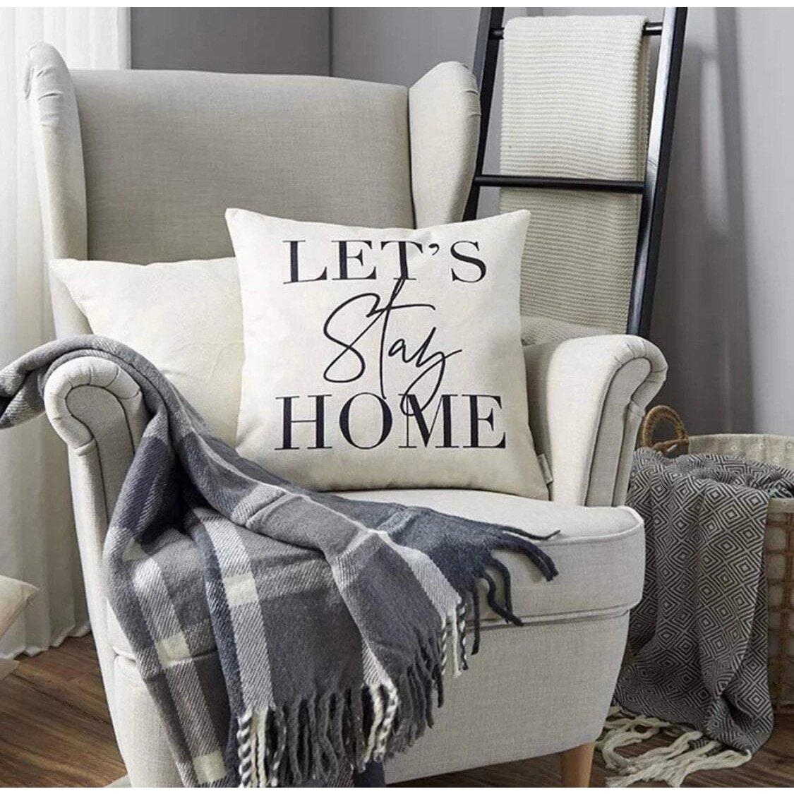 Cream throw cushion with black 'Lets Stay Home' text, on a grey armchair with a tartan blanket, in a living room setting.