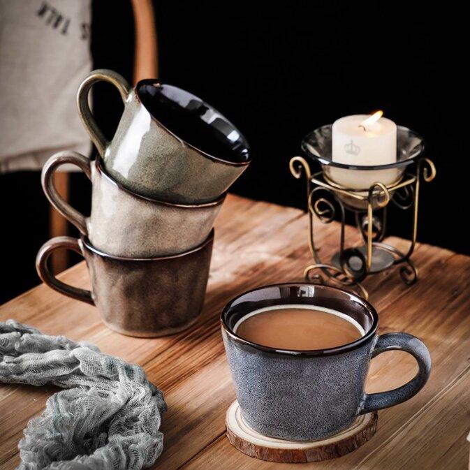 Handmade retro ceramic mugs: three stacked & one with tea on a coaster, on wooden table with a blue material and a lit candle.