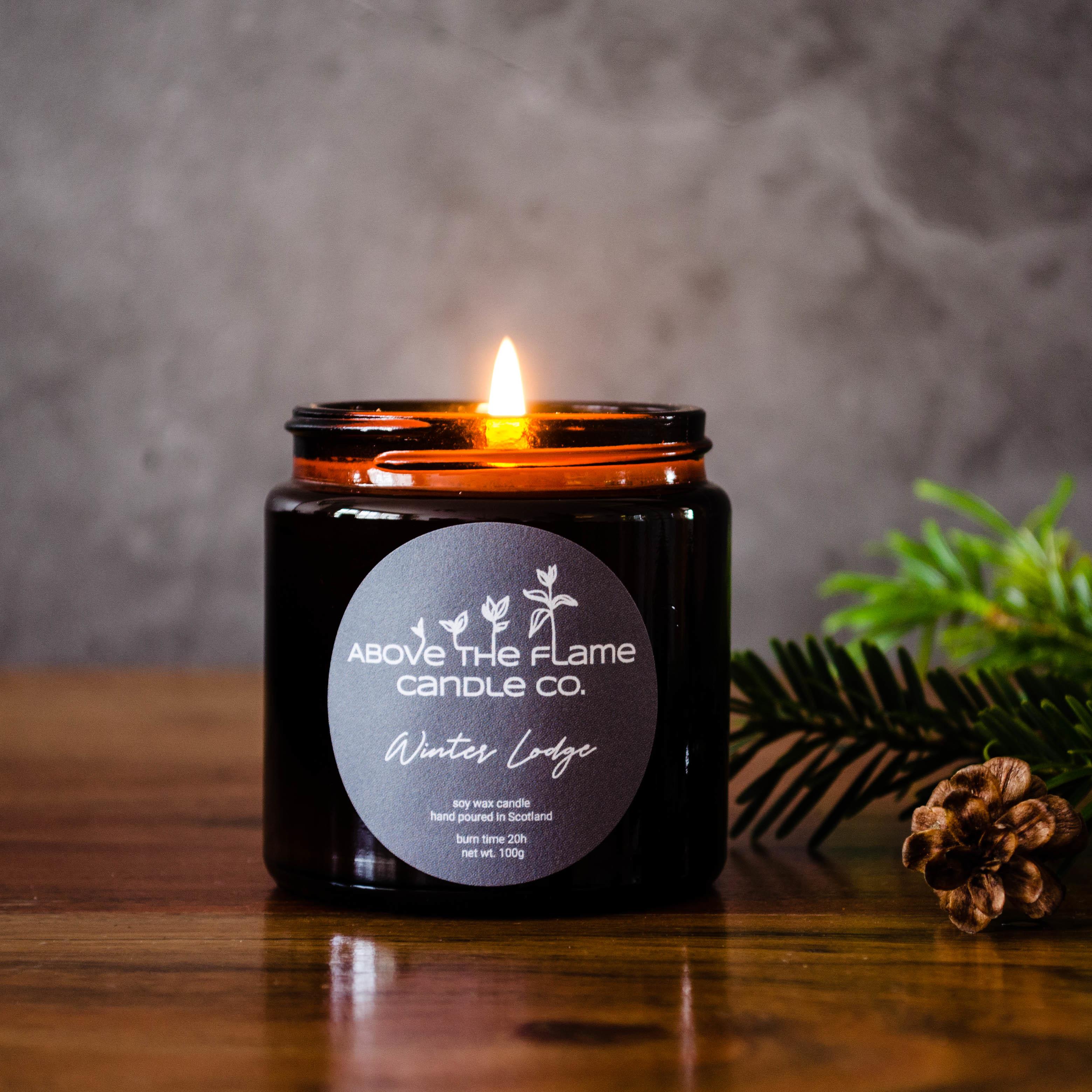 A lit winter lodge amber soy wax candle jar handmade by above the flame candle Co on a wooden table