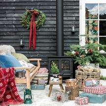 A rustic, cosy outdoor christmas setting with a wooden bench with cushions, a wood burner, blankets, logs, gifts and other decor.