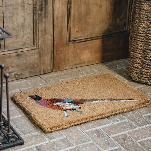 A pheasant doormat on a brick paved step in front of rustic wooden doors, between a boot stand and a wicker basket.