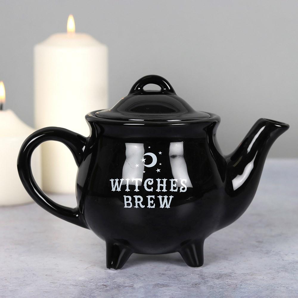 Black ceramic teapot with 'Witches Brew' text and a stars and moon design, on a grey surface in front of lit pillar candles.
