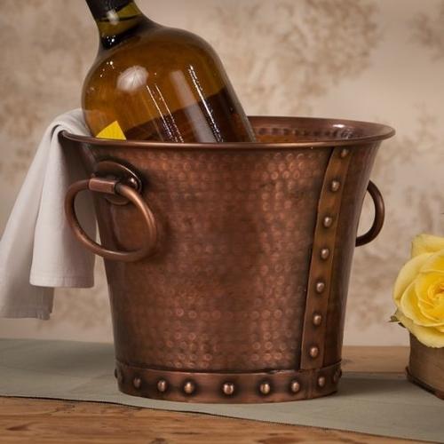 A copper ice bucket on a wooden surface, containing a glass bottle and a white cloth, next to a yellow rose.