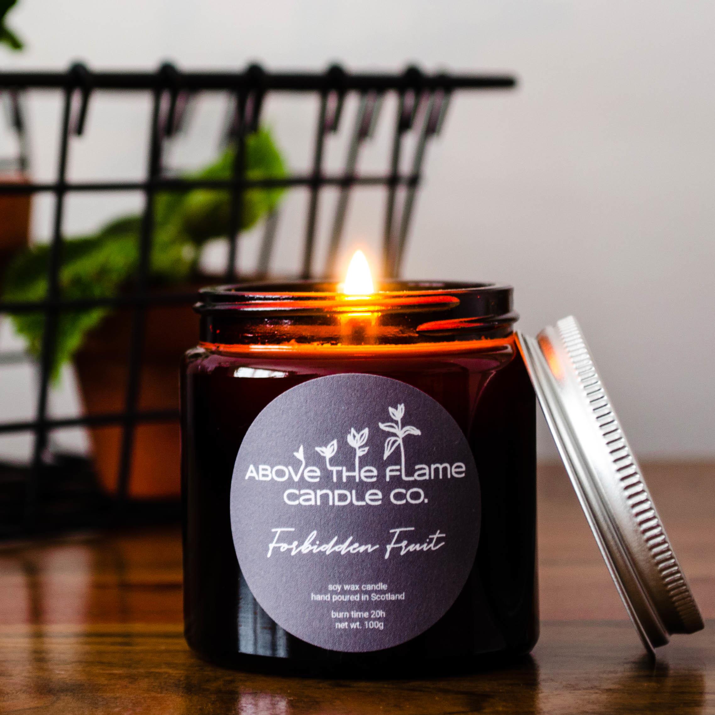 A lit forbidden fruit amber soy wax candle jar handmade by above the flame candle Co on a wooden table