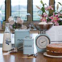 Country kitchen with vintage blue scales, flour & sugar canisters on the worktop, a glass bottle of milk & a victoria sponge.