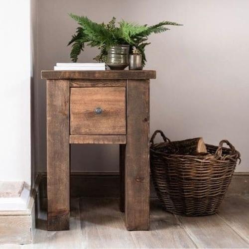 Dark wood slim side table with single square drawer and green plant on top, next to a wicker basket of logs.