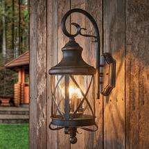 A rustic bronze wrought iron outdoor wall lantern with two bright bulbs, mounted on an old wooden shed wall.