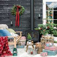 A rustic, cosy outdoor christmas setting with a wooden bench with cushions, a wood burner, blankets, logs, gifts and lanterns.
