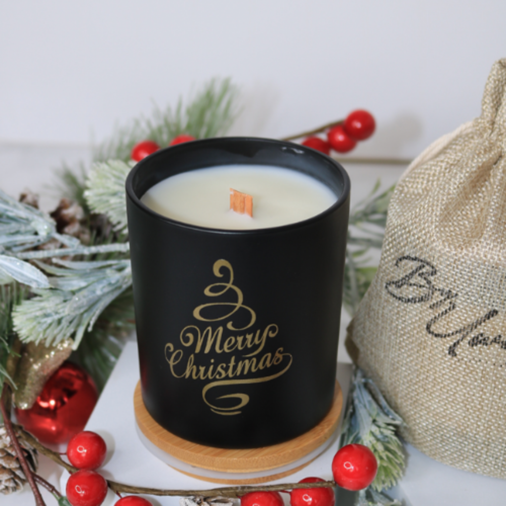 Christmas wish handmade soy wax wood wick candle in black, with gold 'Merry Christmas' text, surrounded by festive foliage.