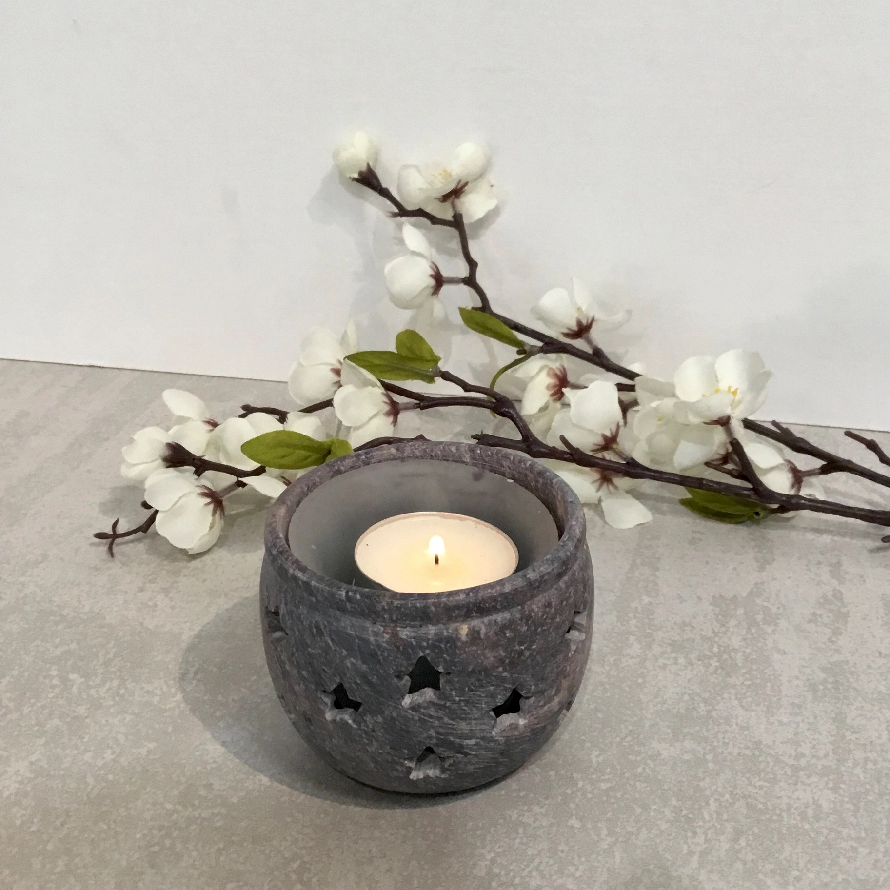 Stone votive tealight candle holder with star cutout design, with a lit tealight on a grey surface in front of white flowers.