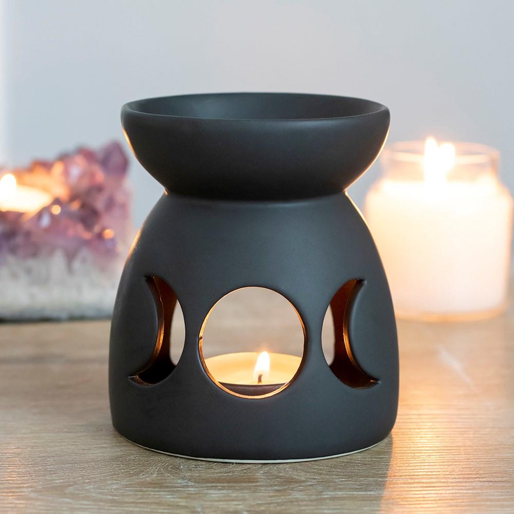 A black ceramic oil burner featuring a cutout triple moon design, shown on a wooden surface with two similar products behind.