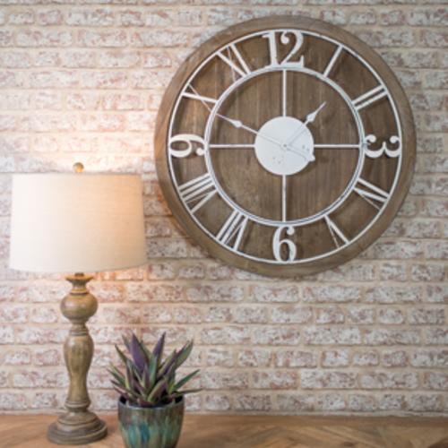 A large rustic wooden wall clock with white roman numerals and hands, on a wall above a wooden sideboard, a lamp and a plant.