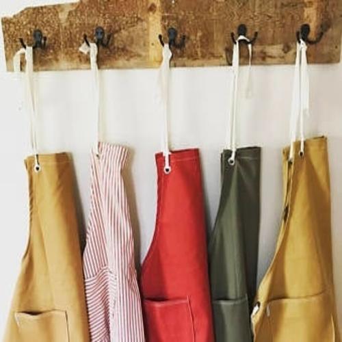 Five coloured aprons hung on a rustic wooden coat hanger board each apron is hung on a seperate block hook.