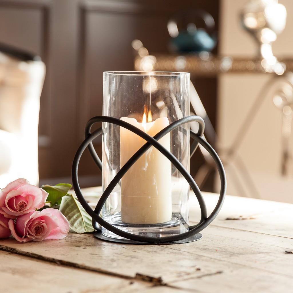 Large metal and glass orbits hurricane candle holder holding a lit pillar candle, on a wooden table with pink roses.