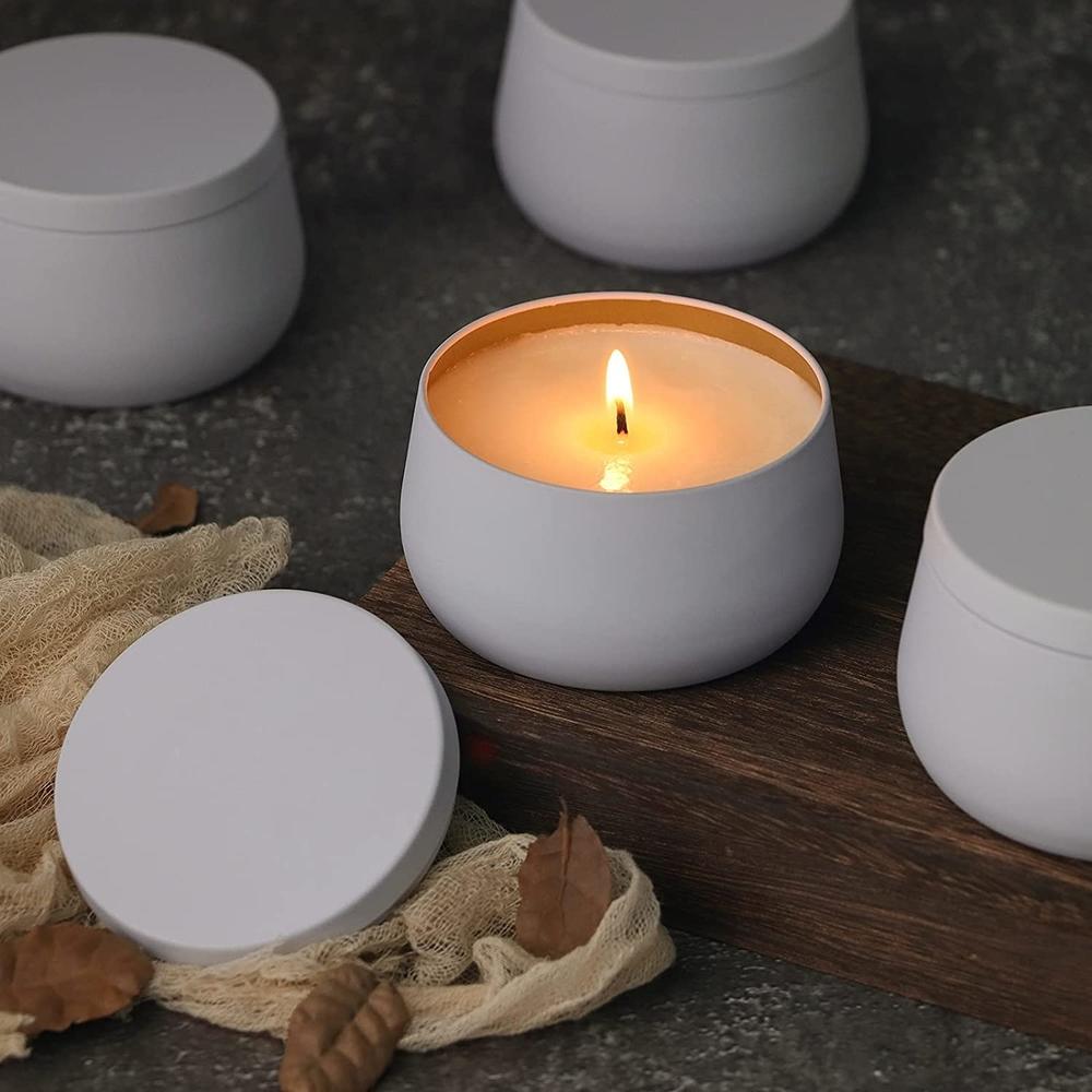 Four white candle tints with one open and lit, on a wooden block and grey surface with brown leaves.