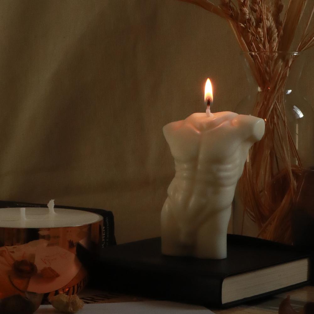 A lit organic soy wax male torso candle, on a black book, next to a round copper candle and wheat plant in a vase.
