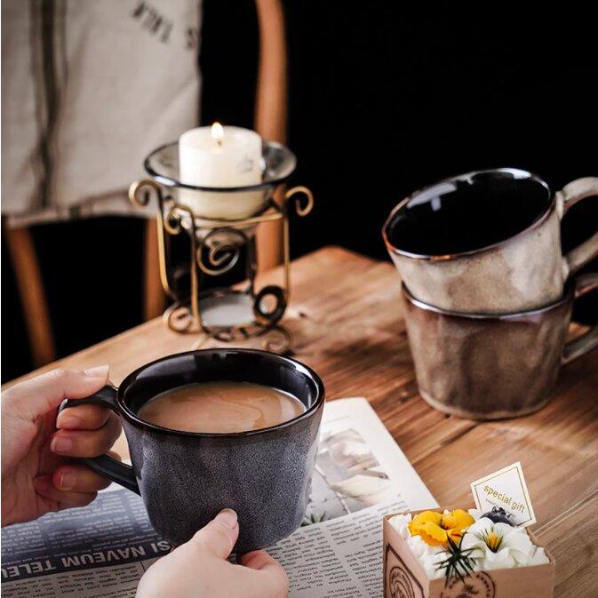 Handmade retro ceramic mugs, two stacked and one containing tea being held, on a wooden table with a candle and other items.