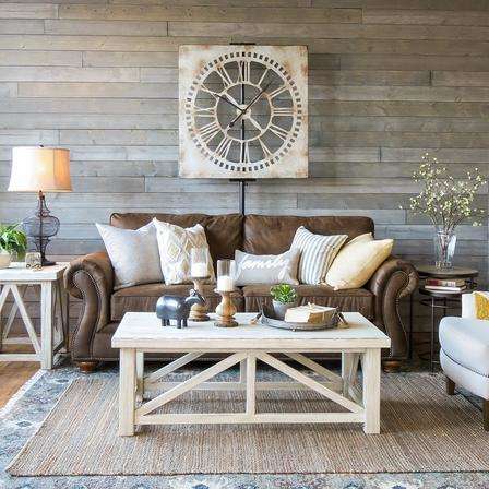 A farmhouse style living room, brown leather sofa with light cushions, square rustic wall clock, white tables and other decor.
