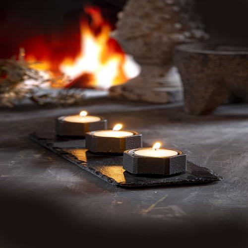 A slate tealight candle holder with three lit tealight candles, sat on the floor in front of an open fire.
