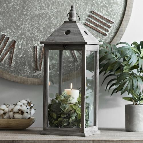 A rustic grey lantern holding a lit pillar candle wrapped in green leaves, sat on a sideboard with a pot plant and potpourri.