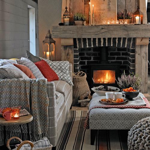 A cosy living room setting with a lit fireplace, a cushion-covered striped sofa, grey ottoman, lanterns & other accessories.