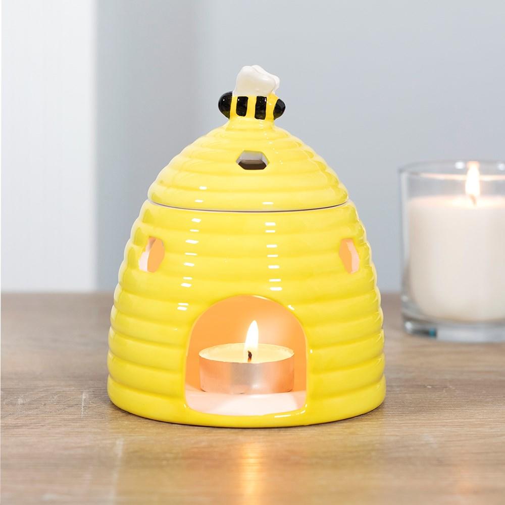 Yellow ceramic oil burner & wax warmer in shape of a beehive with a bee on the lid, on a wooden surface with a candle behind.