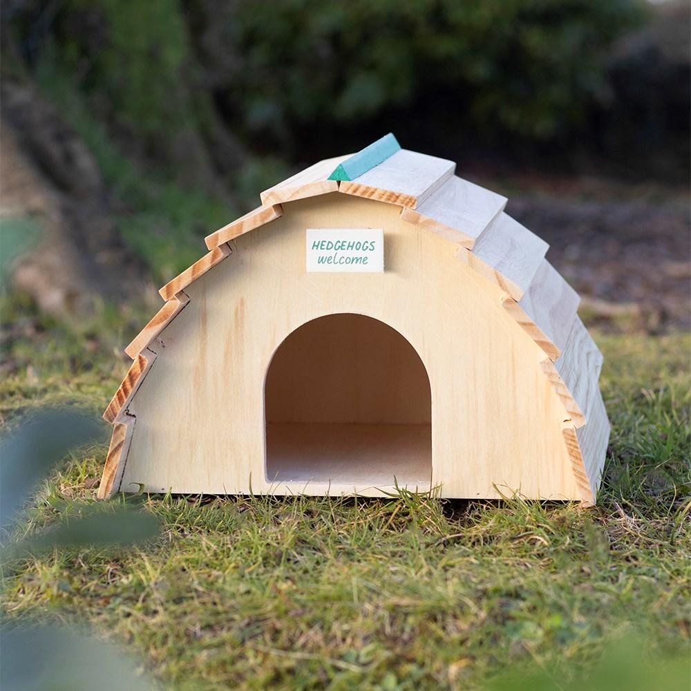 Wooden semi-circle hedgehog house, featuring a sign that reads 'Hedgehogs Welcome' and an opening, shown on grass.