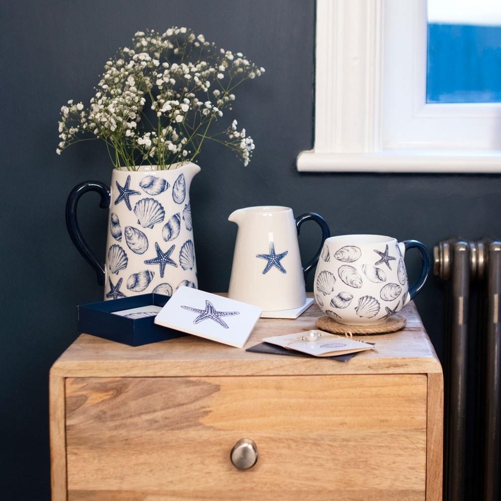 A range of navy and white coastal themed home decor items, including a jug, vase, mug and coasters, on a wooden surface.