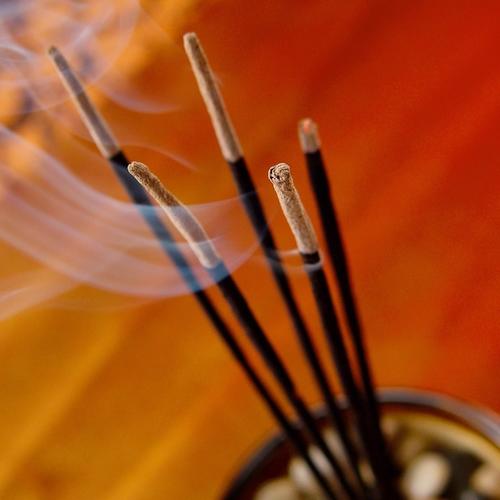 Five lit incense sticks standing up out of an incense holder with the smoke floating across an orange backround.