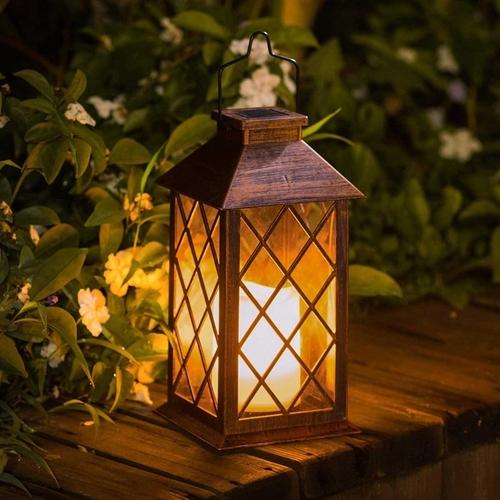 A bronze garden lantern with tudor style glass, holding a lit pillar candle and sat on rustic dark wood in front of flowers.