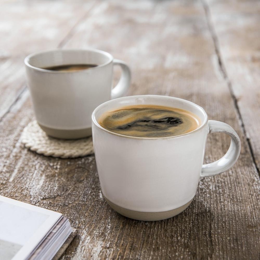 Two white mugs of black coffee, one on a coaster and both on a wooden surface, next to the corner of an open book.