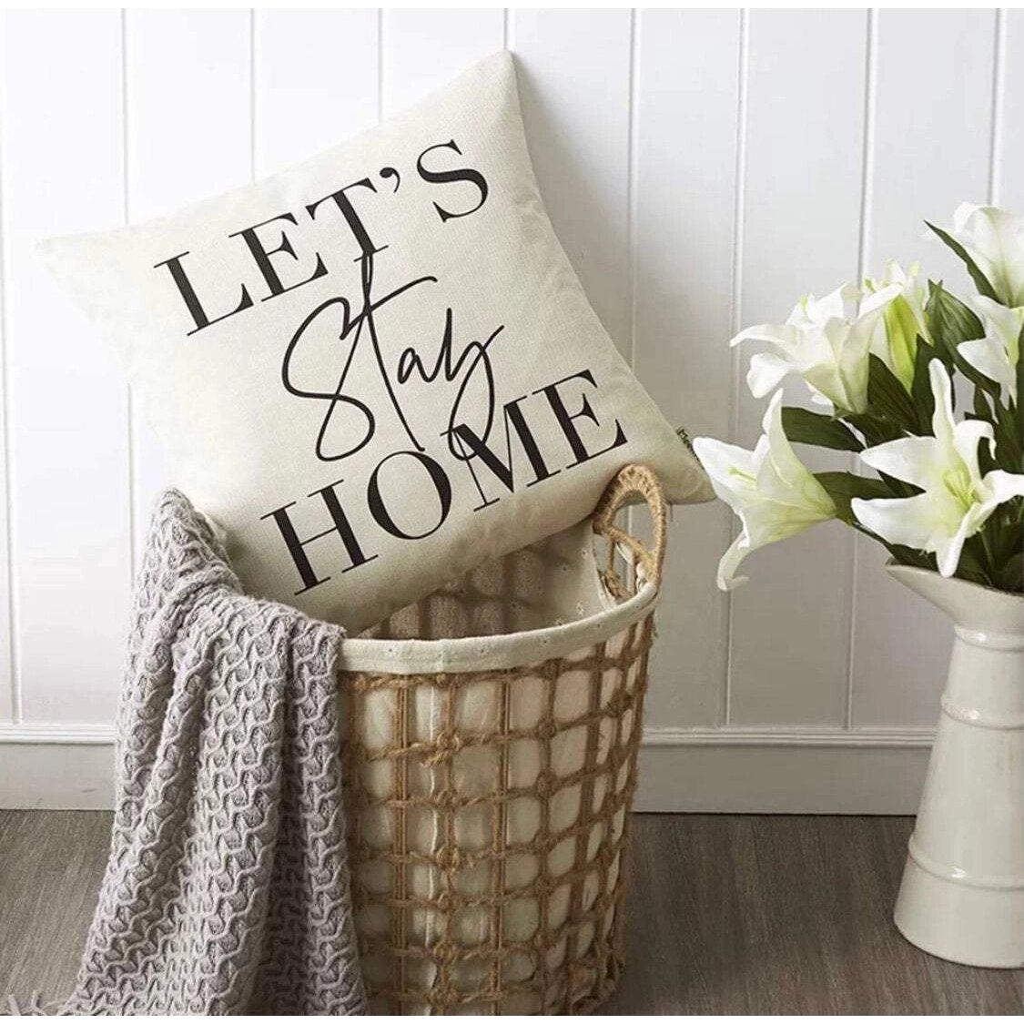 Cream throw cushion with black 'Lets Stay Home' text, on a washing basket with a grey blanket and next to white lilies.
