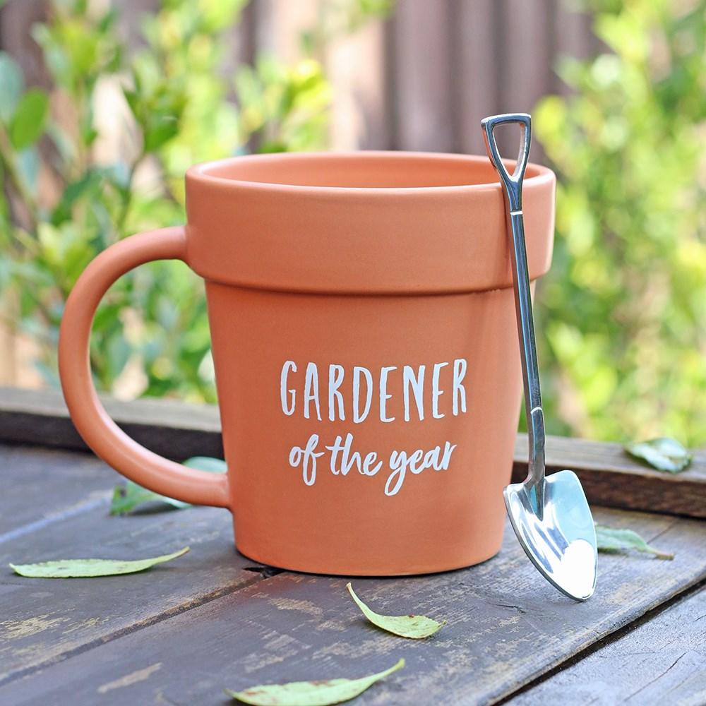 Plant pot-shaped terracotta mug with matching shovel spoon and 'Gardener of the year' text, sat on an outside garden table.