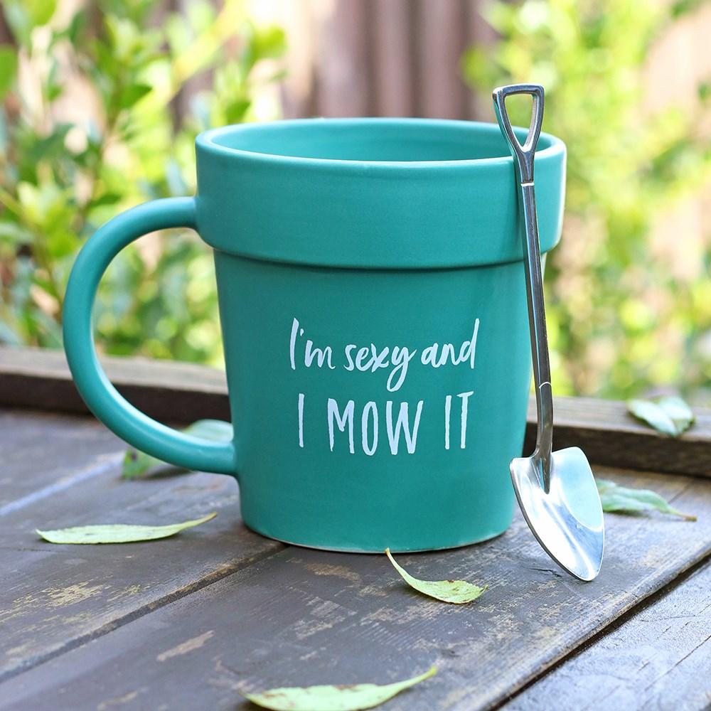 Plant pot-shaped green mug with matching shovel spoon and 'I'm sexy and I mow it' text, sat on an outside table with leaves.