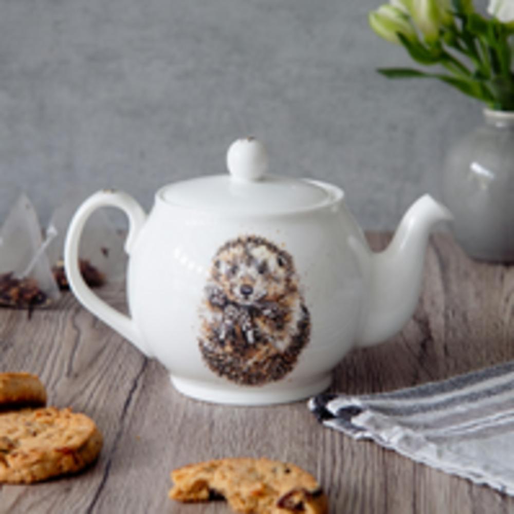 White ceramic teapot with hedgehog design, on a wooden surface surrounded by cookies, a potted plant and a tea towel.