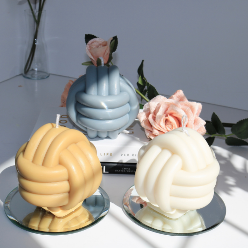 Three giant knot handmade Soy Wax Candles: blue, yellow, and white, on a white surface with pink roses.