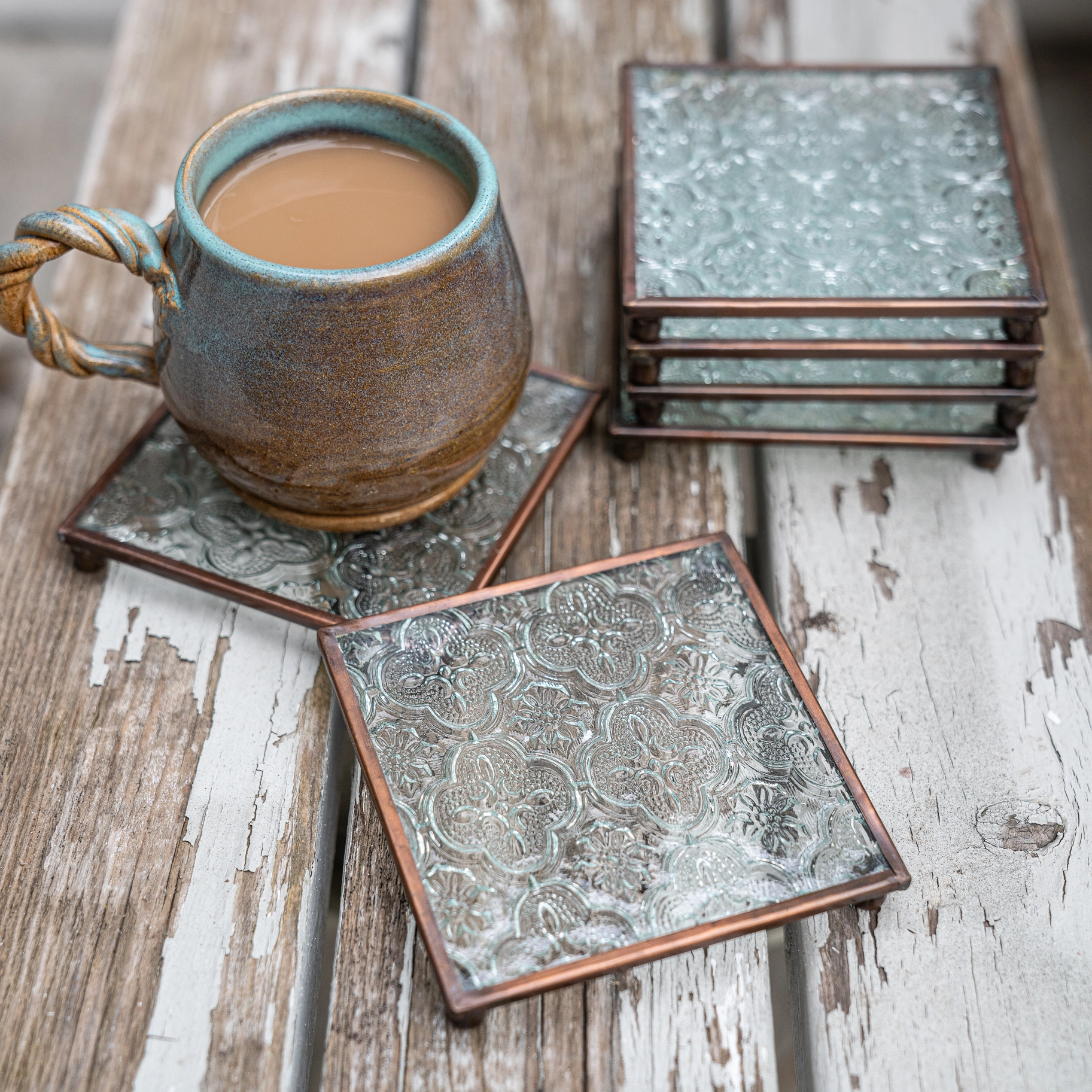 Six patterned copper and glass pillar plates, four stacked and one holding a mug of tea on a wooden bench.