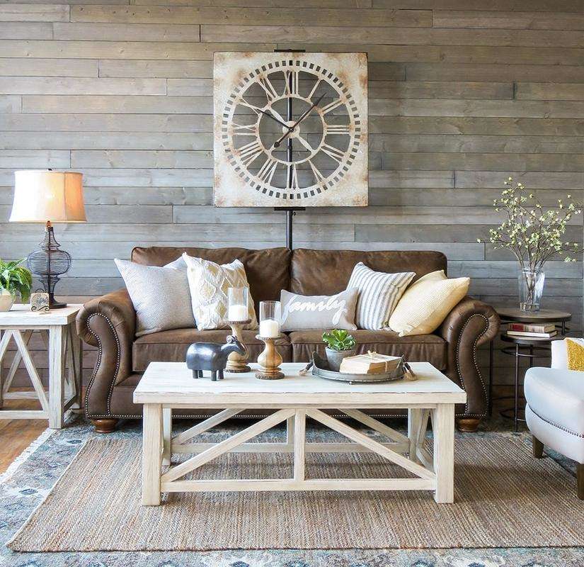 A farmhouse style living room, brown leather sofa with light cushions, square rustic wall clock, white table and other decor.