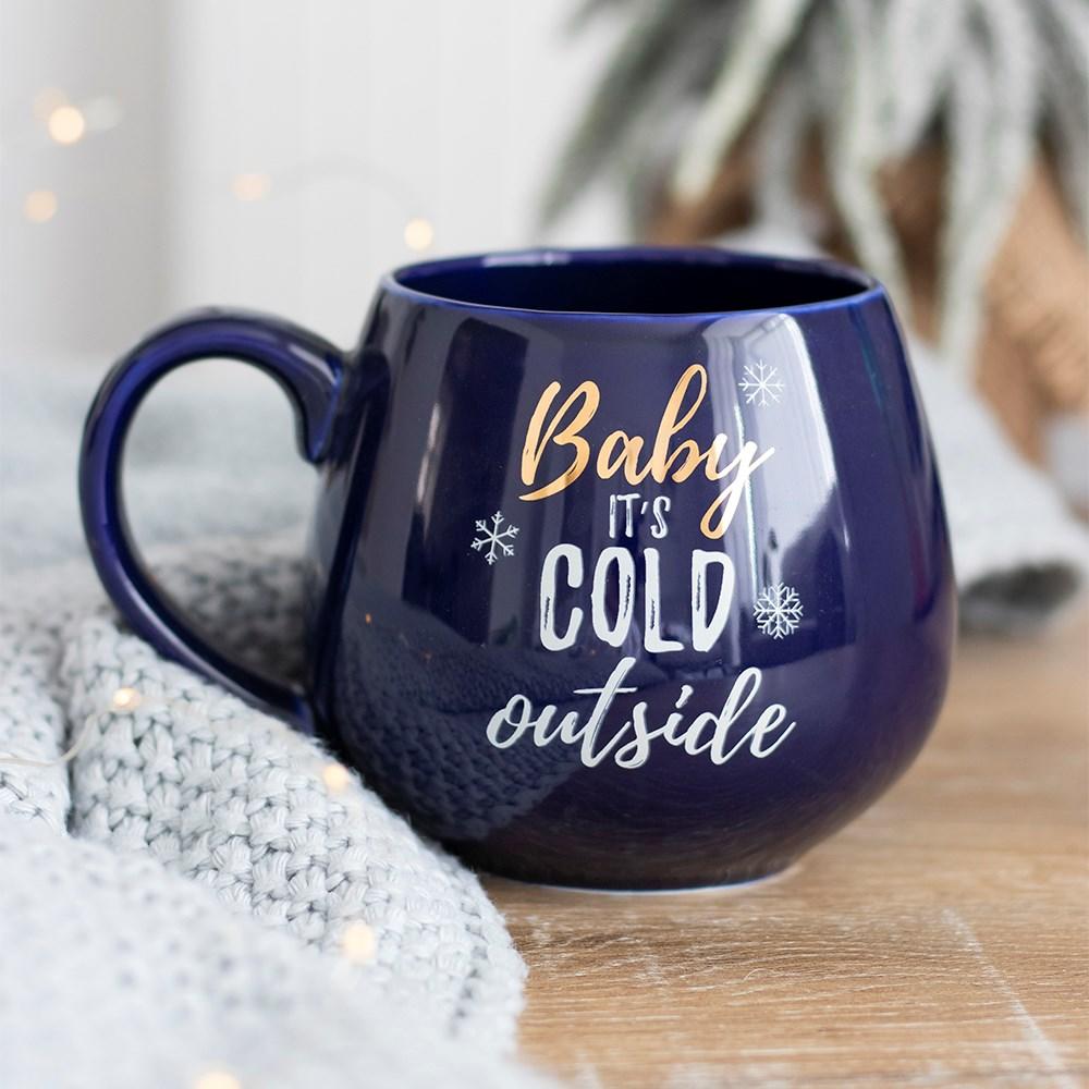 Navy Christmas mug with gold and white 'Baby its cold outside' text and snowflakes, on a wooden surface with a grey blanket.