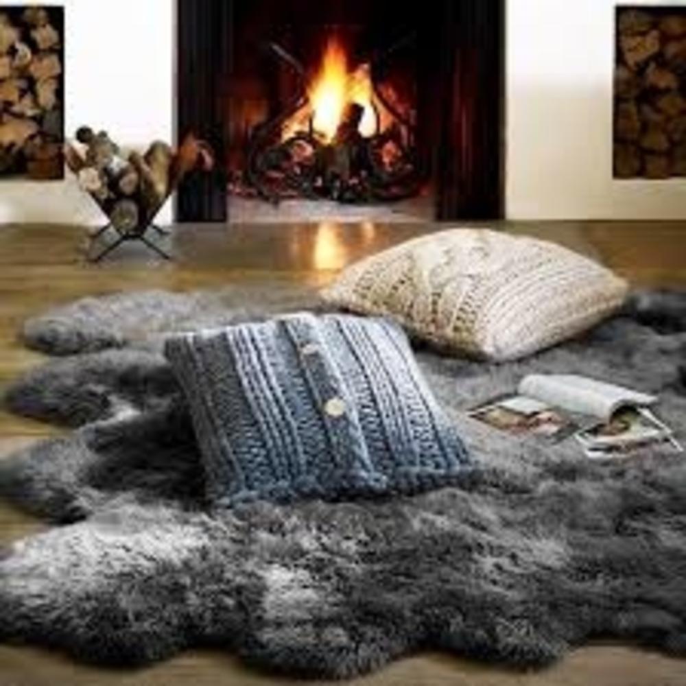 Grey sheepskin rug, two knitted pillows and magazines in front of a lit fireplace and logs.
