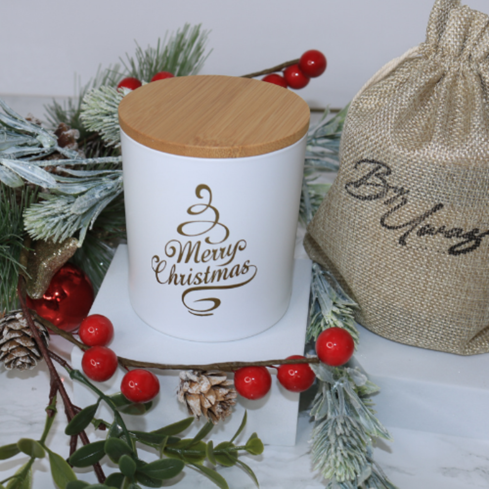 Christmas wish handmade soy wax wood wick candle in white, with gold 'Merry Christmas' text, surrounded by festive foliage.