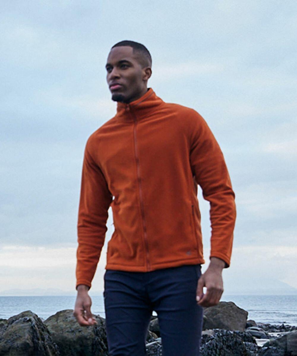 A person wearing an orange Craghoppers CR315 jacket and black pants stands near a rocky shore under a cloudy sky.