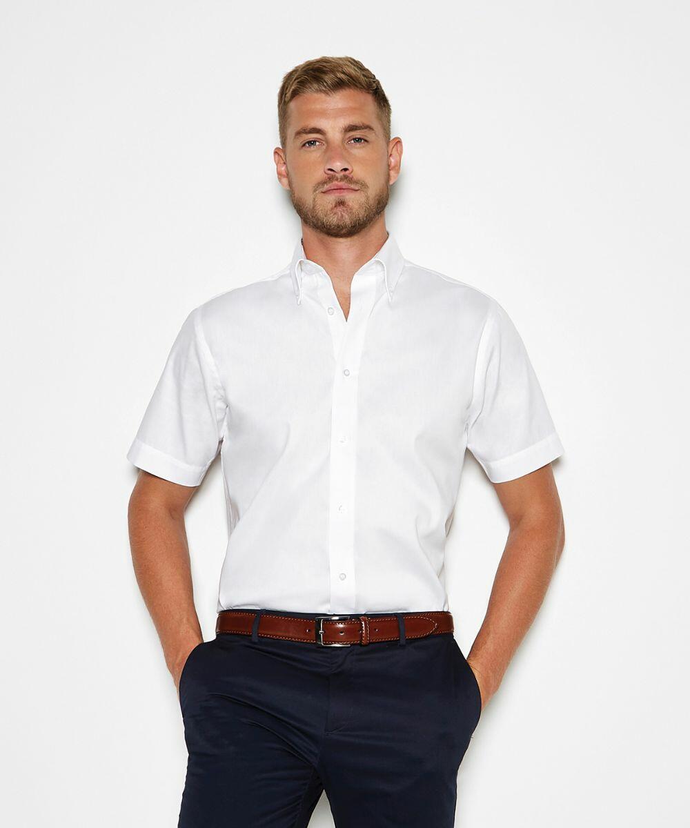 A man with short hair wears a white Kustom Kit KK187 short-sleeve button-up shirt and dark trousers, posing against a plain white background with hands in pockets.
