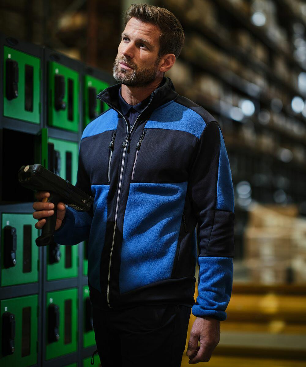 A man wearing a blue and black Regatta RG545 jacket holds a scanner in a warehouse, with green lockers in the background.