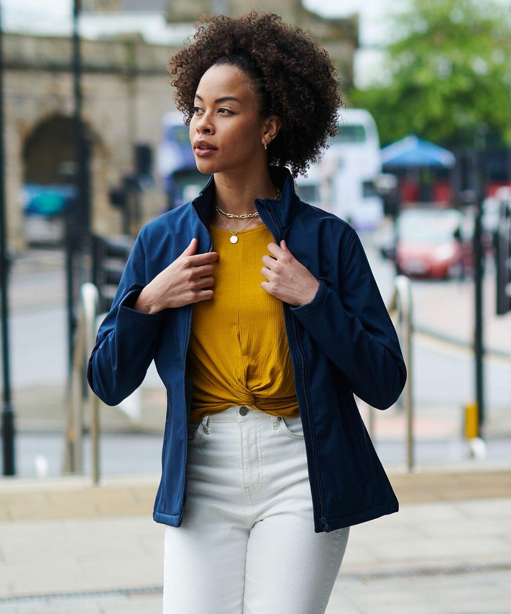 A woman walks outside wearing a Regatta RG027 jacket over a yellow top, paired with white pants. She has curly hair and holds the jacket collar with both hands. The background includes buildings and parked cars.